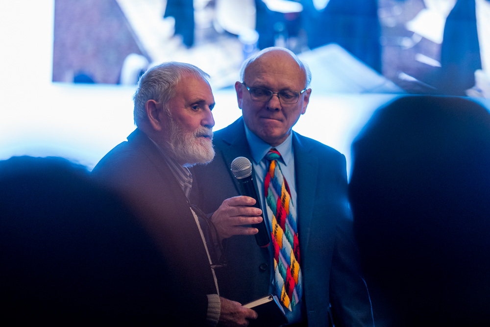David Wallace speaks into a microphone while Ron Hauenstein stands beside him in the audience.