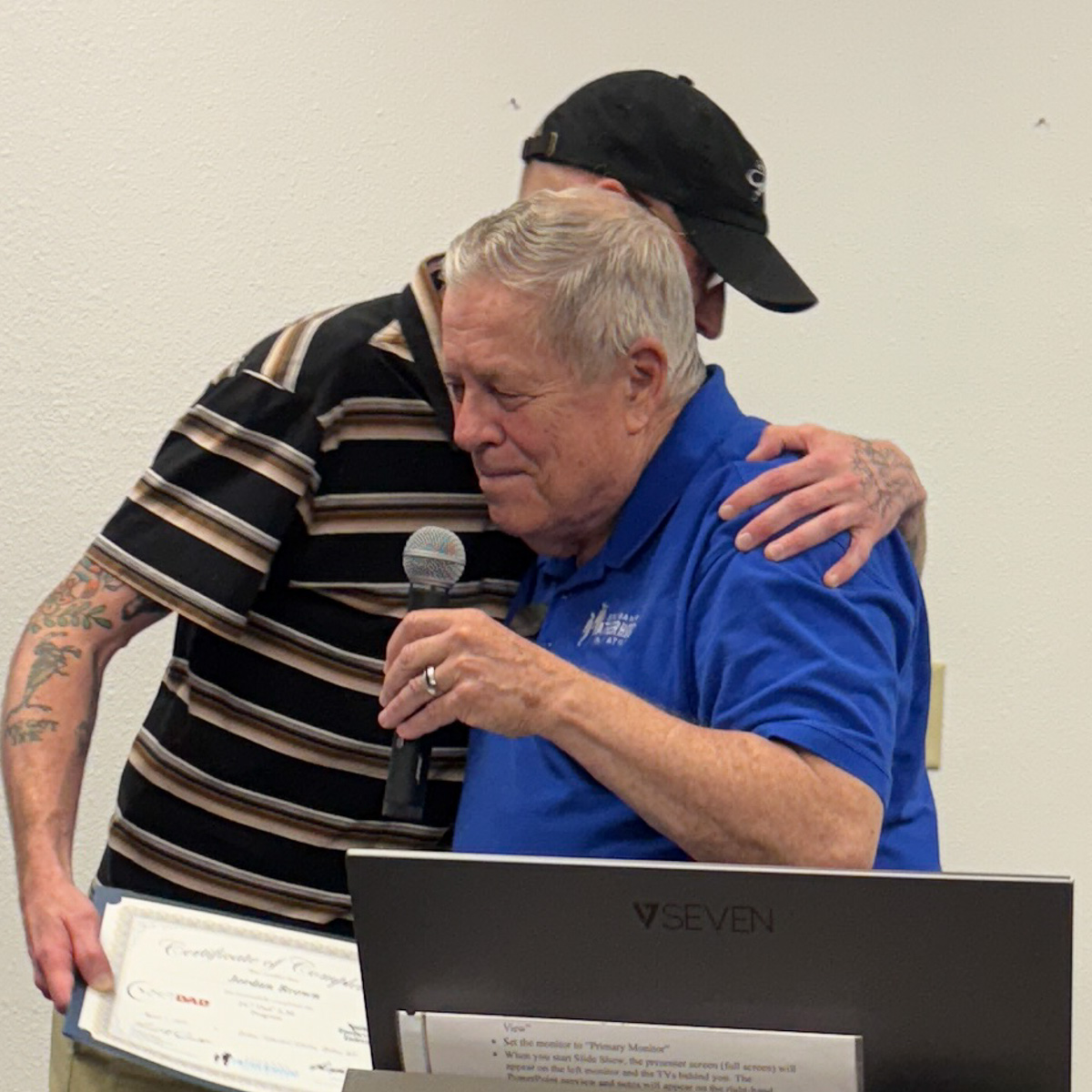 Tattooed father in striped shirt embracing instructor in blue SpoFI shirt during graduation ceremony