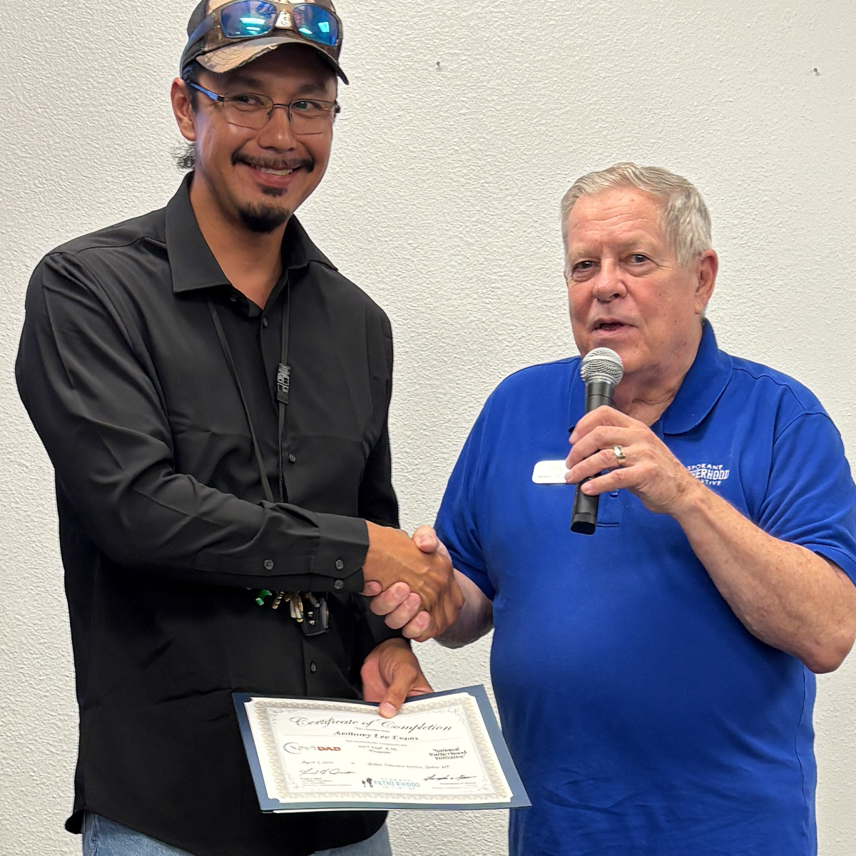 Father in dark business shirt shaking hands with instructor while holding graduation certificate