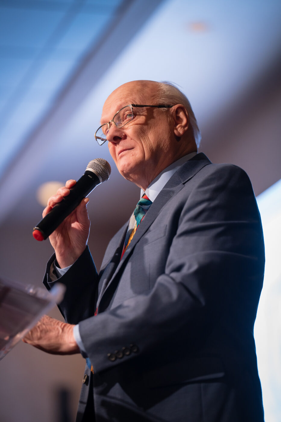 Ron Hauenstein speaks with conviction into a handheld microphone on stage at the SpoFI 10th anniversary celebration at Mirabeau Park Hotel.