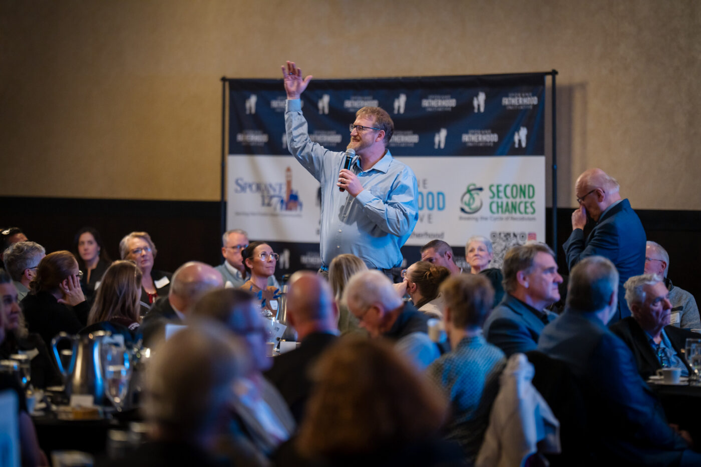 Nathan Henry stands with hand raised and speaks into a microphone from the audience at the SpoFI 10th anniversary celebration, with SpoFI, Spokane127 and Second Chances banners visible behind him.
