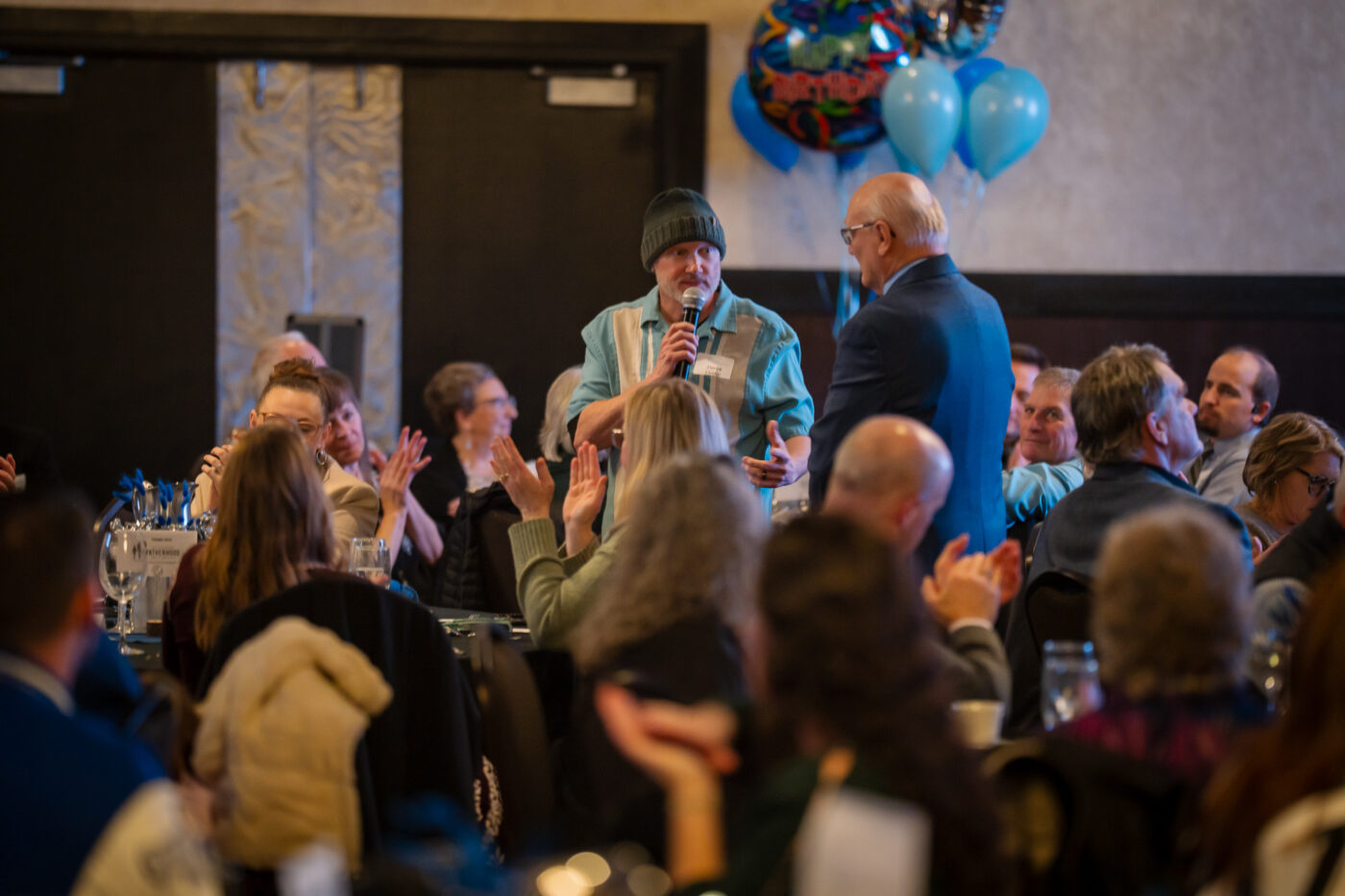 Derek Cutlip of Shine FM stands and speaks into a microphone while the room listens at the SpoFI 10th anniversary celebration.