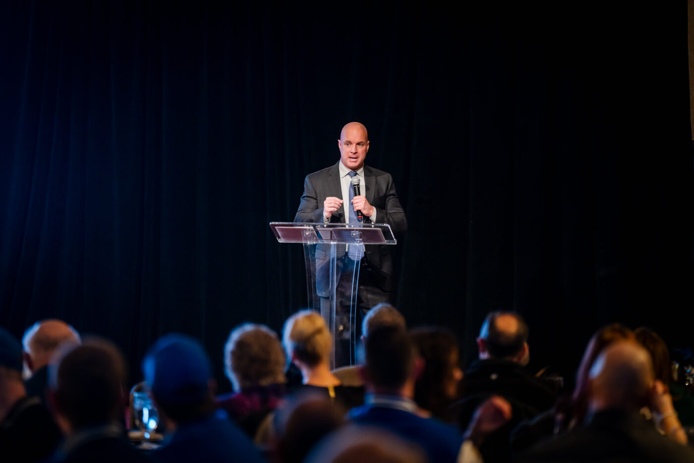 Former Police Chief Craig Meidl delivers the keynote address at a clear podium with dramatic lighting.