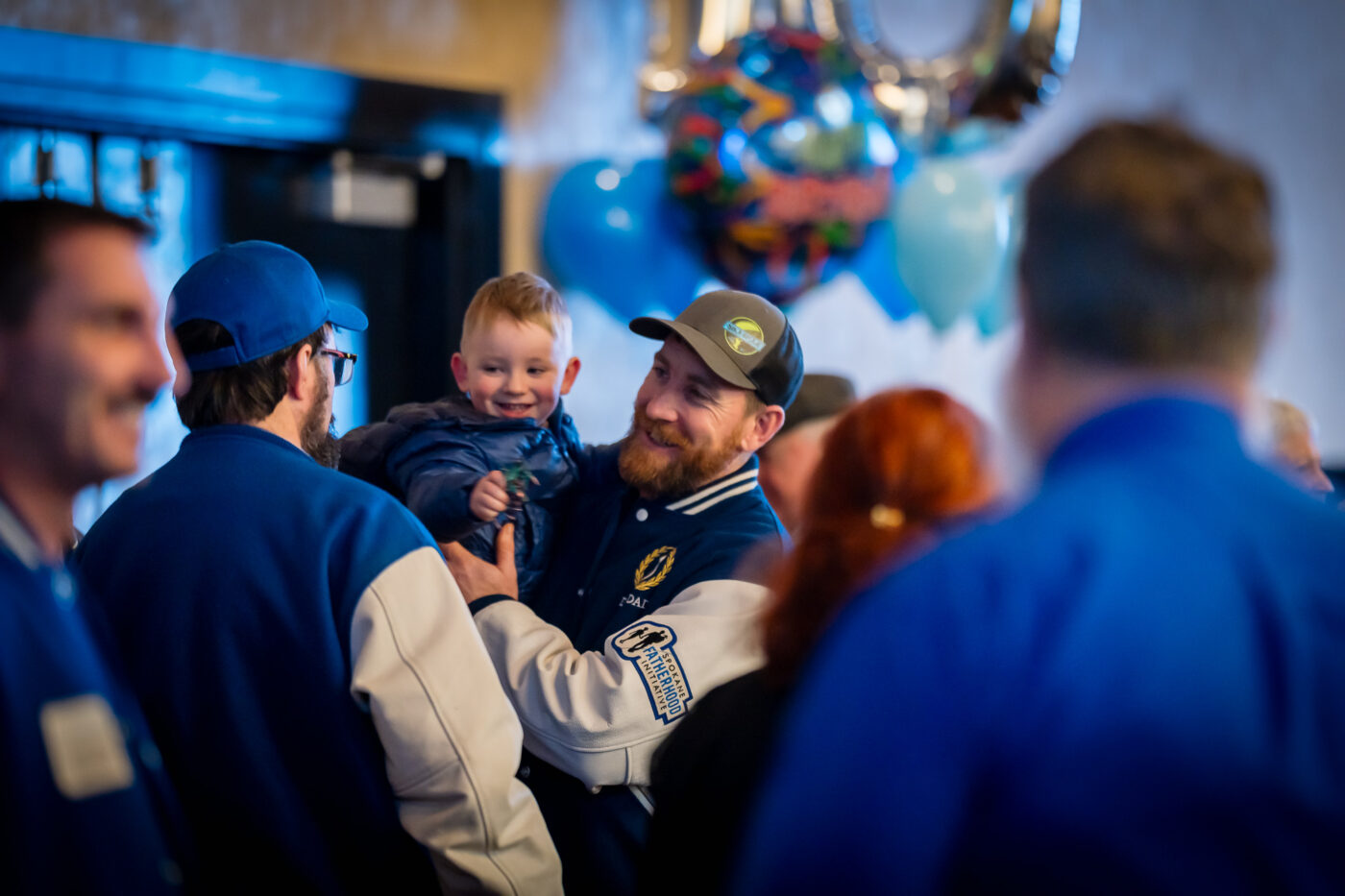 Dad2Dad graduate and facilitator Garrett Skirvinn holds his smiling toddler son surrounded by fellow graduates in their Dad2Dad letterman jackets at the Spokane Fatherhood Initiative 10th anniversary celebration