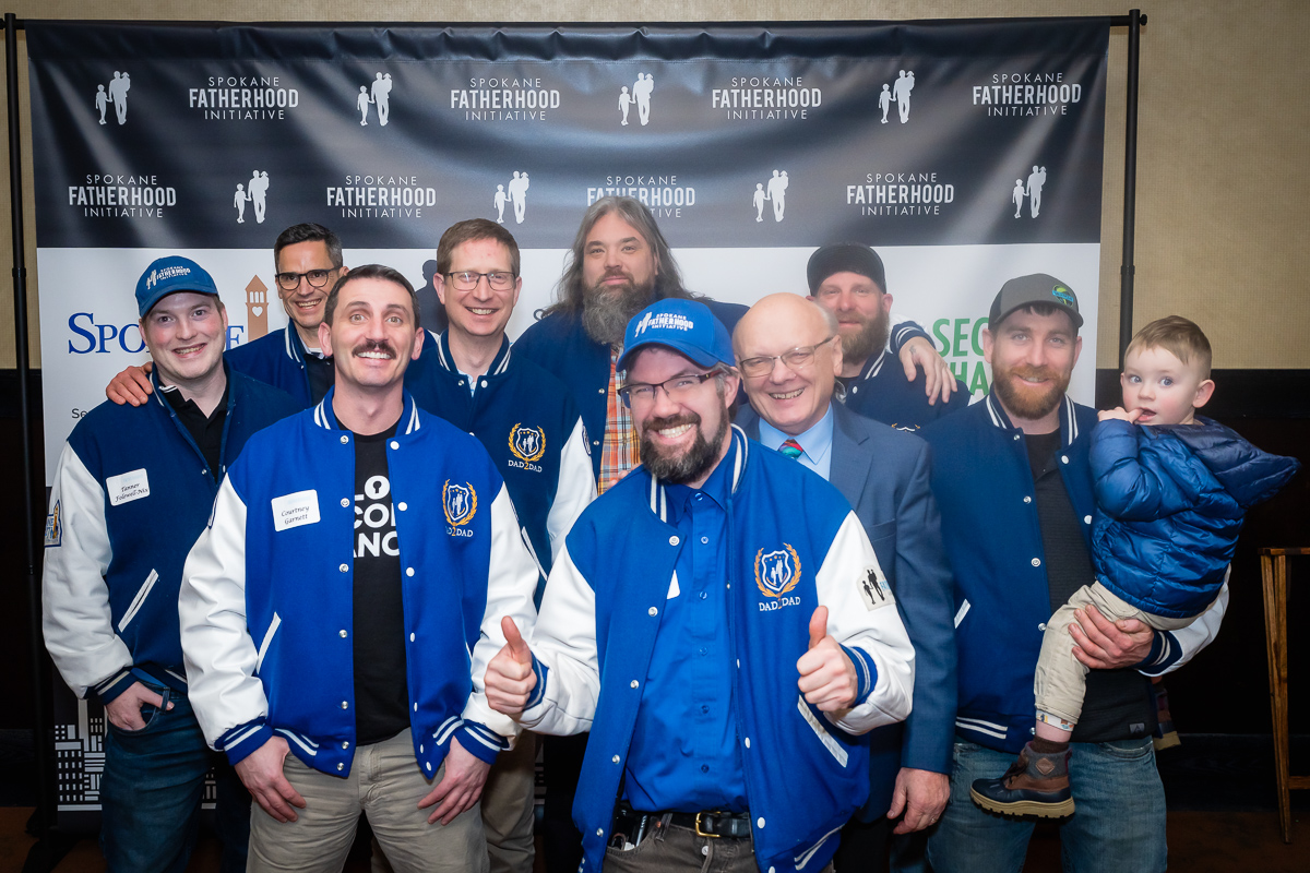 Ron Hauenstein poses with Dad2Dad graduates wearing SpoFI letterman jackets in front of a Spokane Fatherhood Initiative backdrop; one dad holds a toddler.