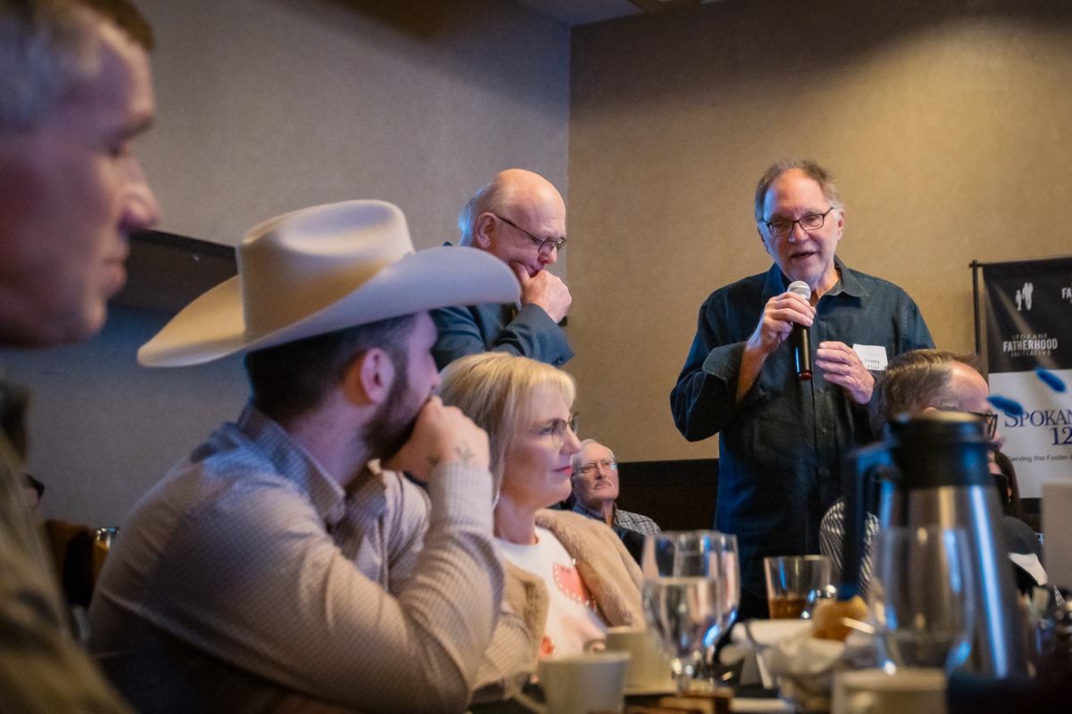 Denny Klaja speaks into a microphone from his table while Ron Hauenstein listens at the SpoFI 10th anniversary celebration.