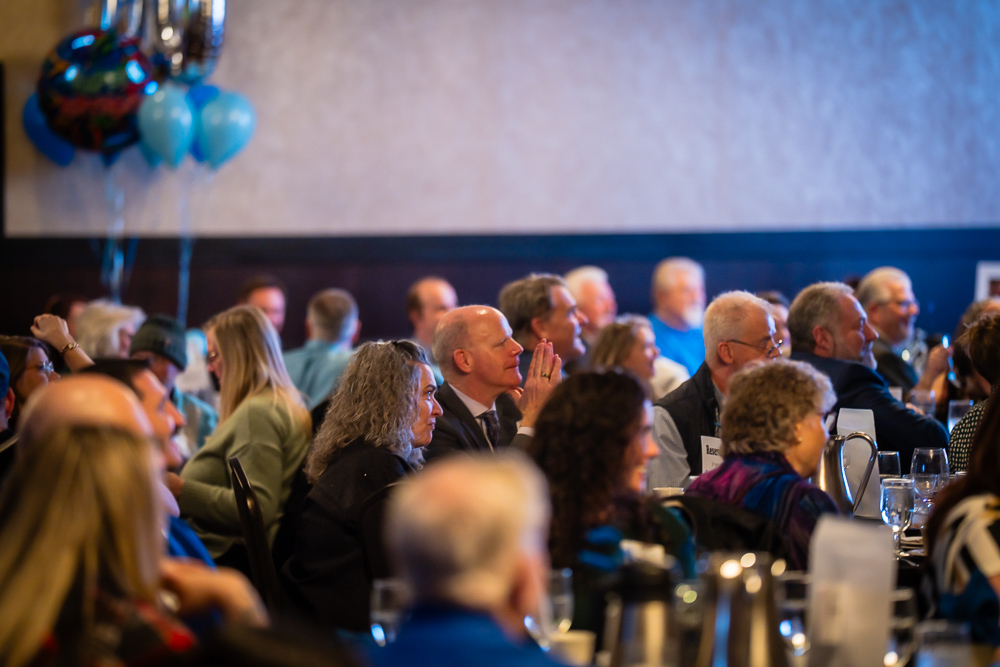 Geoff Swindler seated among banquet attendees, hands raised in applause, listening during the SpoFI 10-year celebration program.