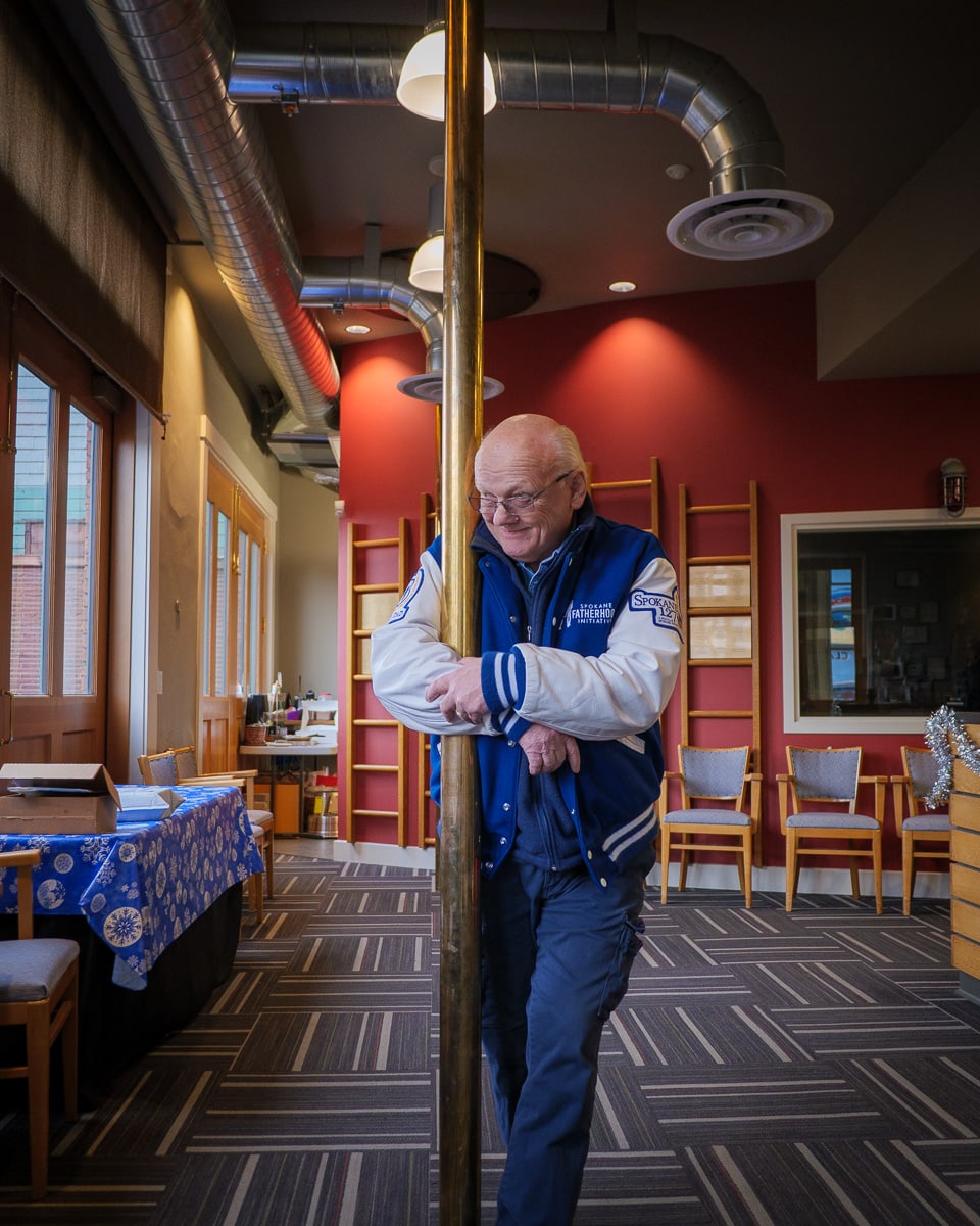 Ron Hauenstein stands at the original brass fireman's pole inside Spokane Public Radio's renovated historic firehouse studios, wearing his SpoFI letterman jacket with exposed industrial ductwork visible above