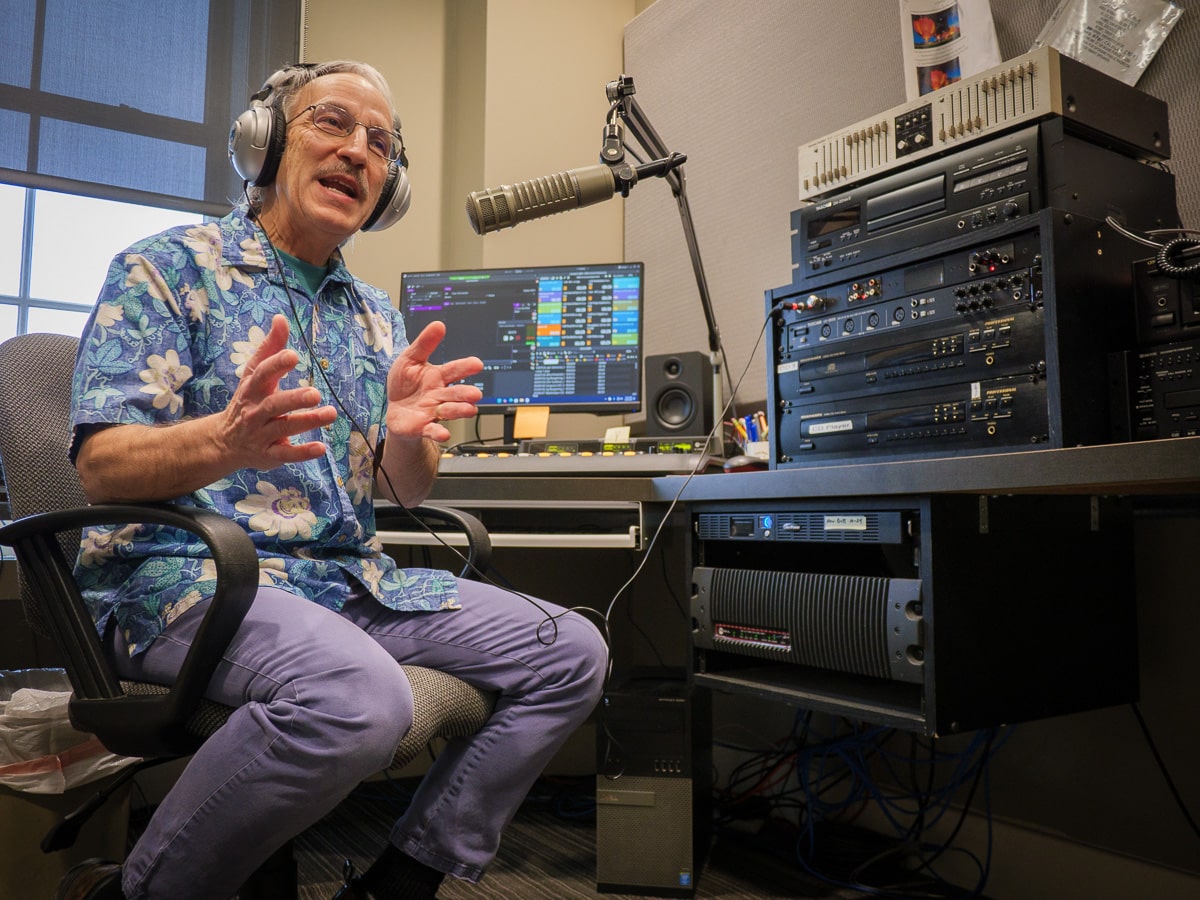 Doug Nadvornick, Spokane Public Radio News Director, speaks animatedly in the broadcast control room wearing headphones and a blue floral shirt, with professional audio equipment and monitors visible behind him