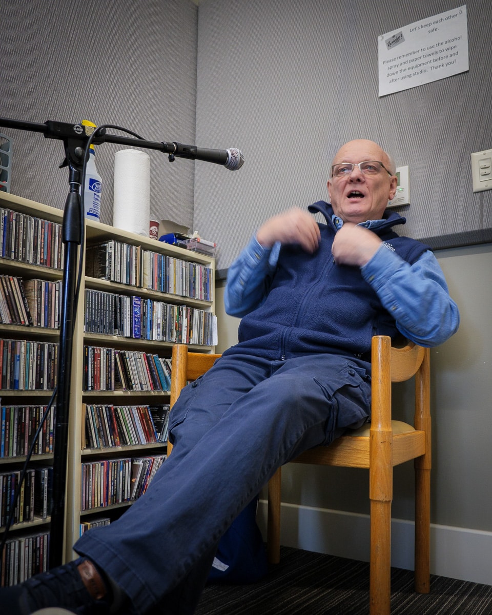 Ron Hauenstein gestures expressively while speaking during the interview, seated in the studio chair wearing his SpoFI fleece, with CD shelves visible behind him