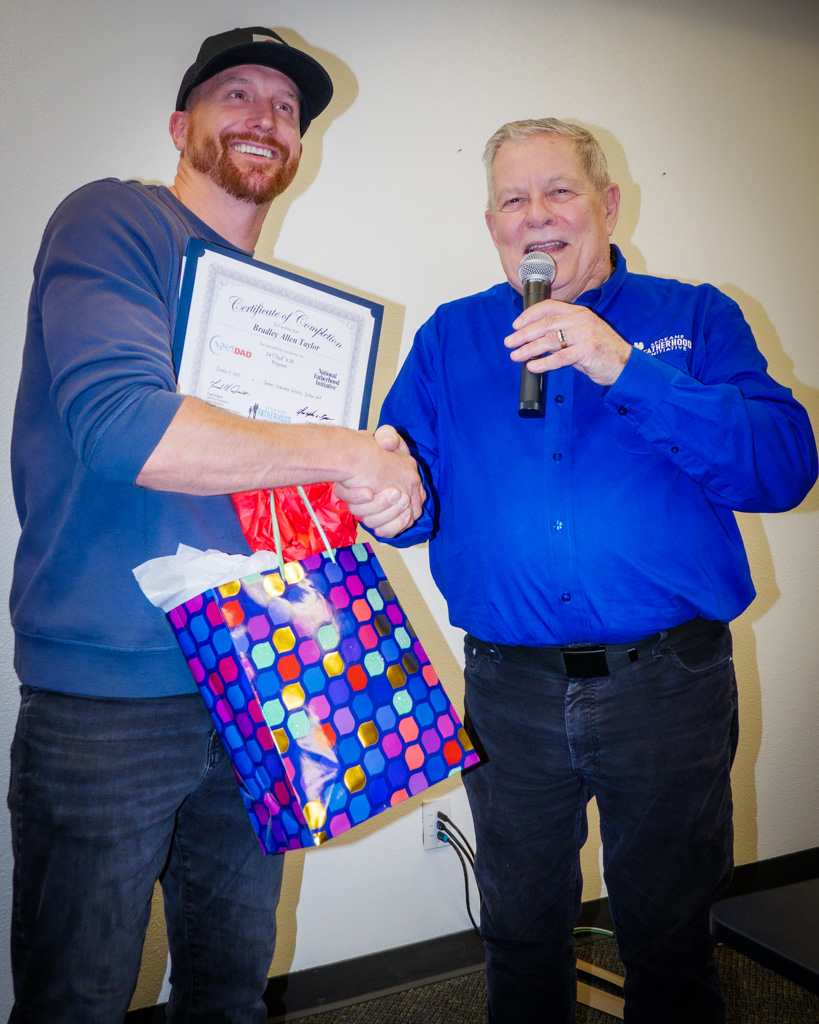 Instructor and SpoFI Vice President Fred Dent speaks into a microphone during a 24/7 Dad® graduation ceremony, addressing fathers and families gathered to celebrate program completion.