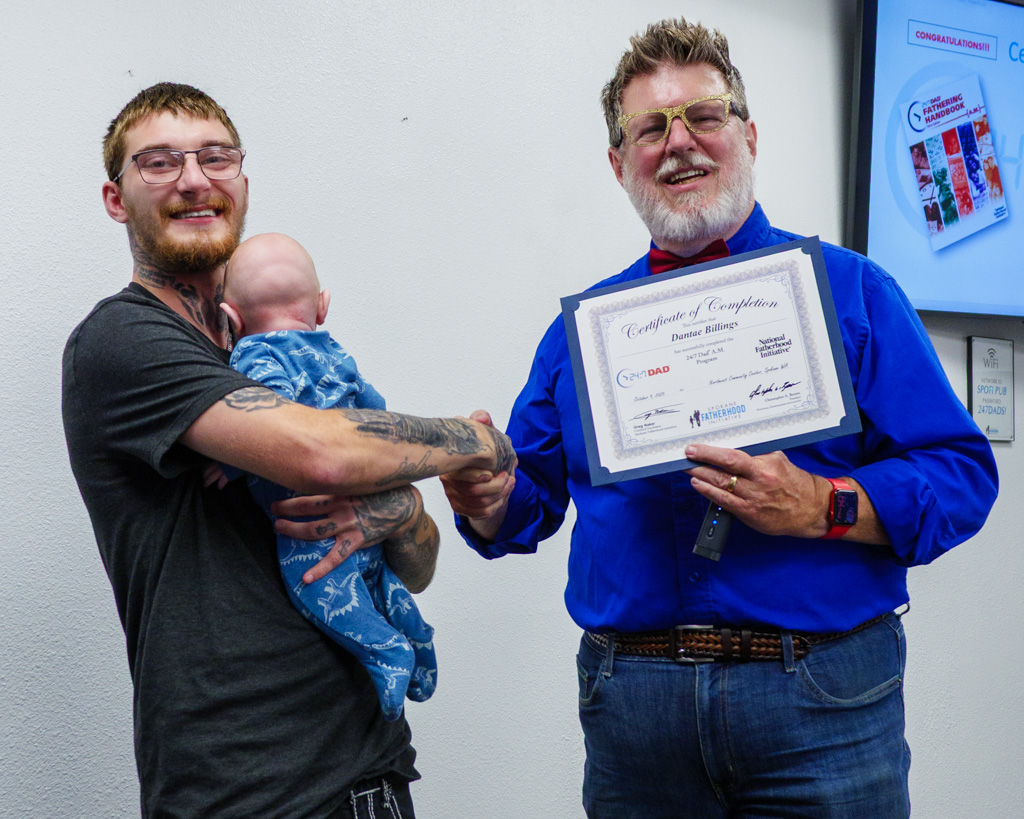 SpoFI instructor Greg Naker presents graduate Dantae Billings with his 24/7 Dad® A.M. Certificate of Completion as Dexter holds his infant son during the October 2025 graduation celebration.