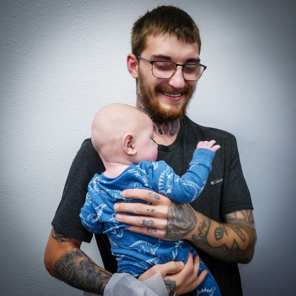 Graduate Dantae Billings smiles while holding his infant son after receiving his 24/7 Dad® A.M. Certificate of Completion during the October 2025 Spokane Fatherhood Initiative graduation.