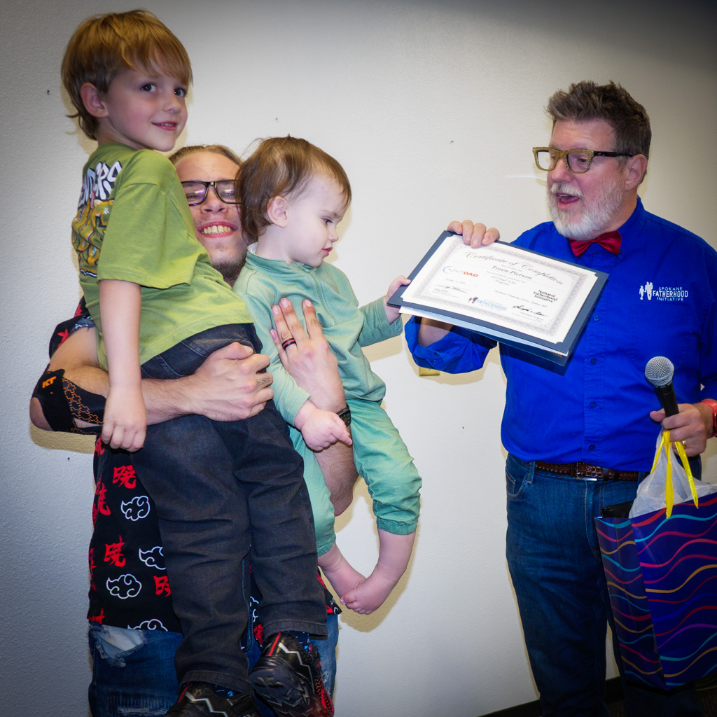Father holding two small children while Greg Naker presents the certificate; all smiling during the ceremony.