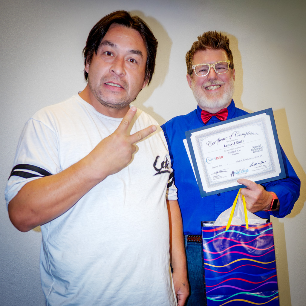 A man in a white T-shirt poses with his certificate next to Greg Naker, who holds a colorful gift bag.