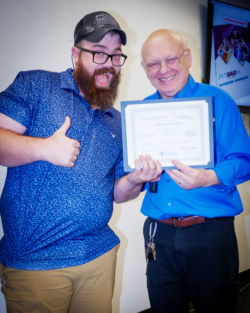 Matt Mellott gives a thumbs-up beside Fred Dent, both holding his 24/7 Dad certificate and smiling broadly.
