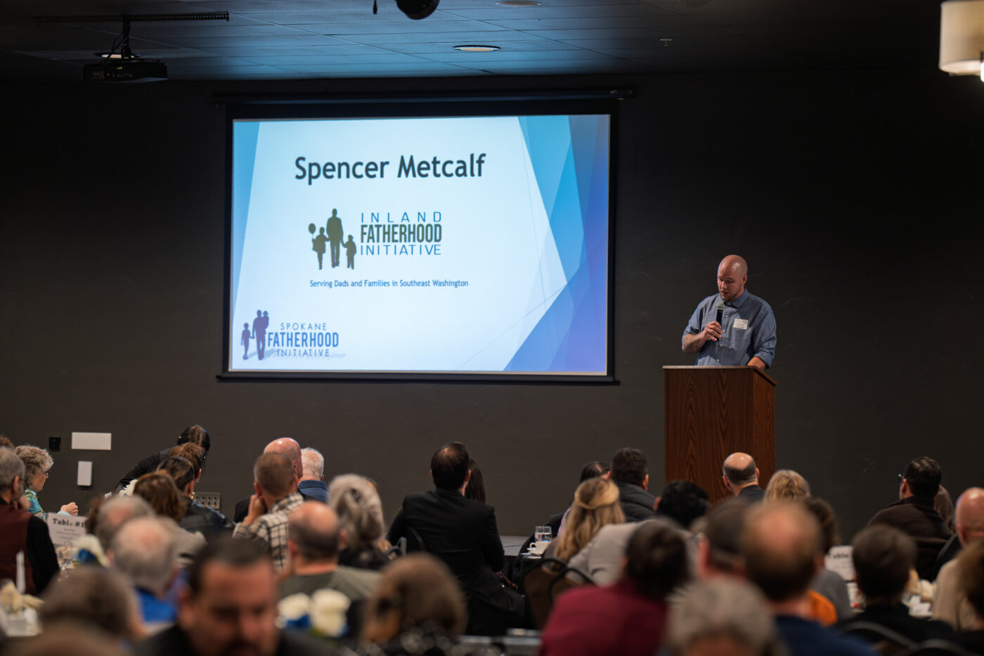 Building Legacy: Nearly 200 Gather to Celebrate Fatherhood and Family Transformation 3 Spencer Metcalf speaks at podium in front of large presentation screen displaying his name and Inland Fatherhood Initiative logo while audience members listen from dinner tables