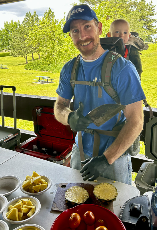 SpoFI Alumni: Step Up and Lead – June 15th Picnic 2 SpoFI alumni father wearing blue program shirt and baseball cap, carrying his baby in a back carrier while volunteering at the juice station during the Father's Day picnic, preparing fresh fruits visible on the serving table