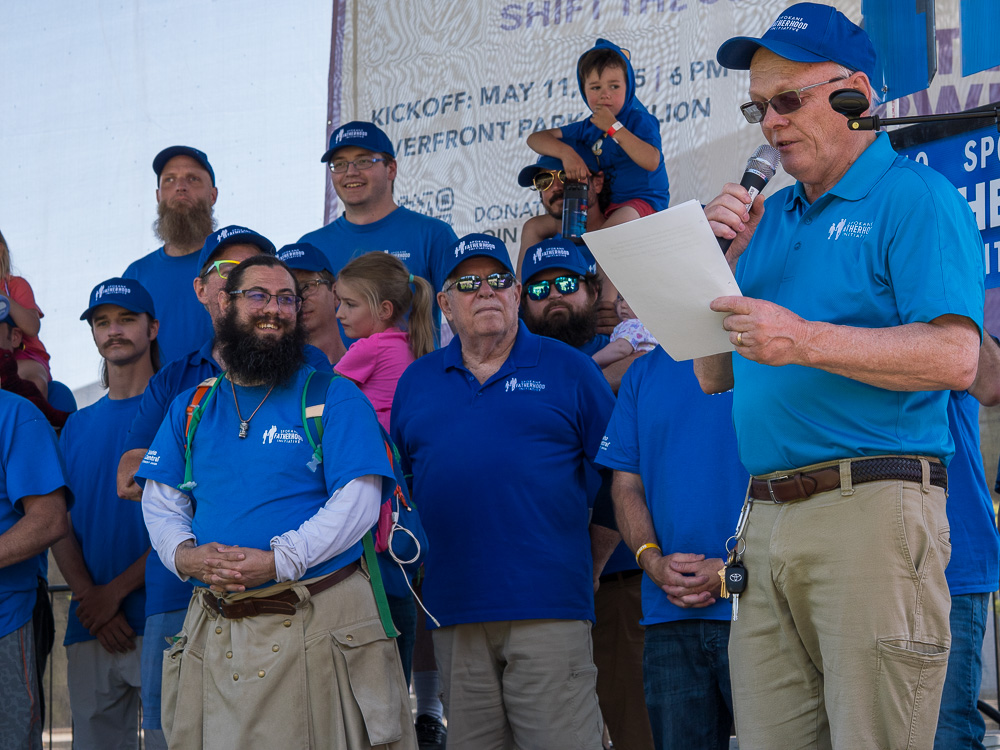 Community connection strengthens Spokane families at Father's Day celebration 10 SpoFI leader speaks into microphone during Father of the Year ceremony as other fathers in blue shirts gather on stage