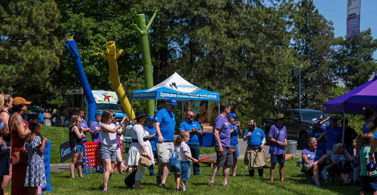 Community connection strengthens Spokane families at Father's Day celebration 4 Wide view of Spokane families enjoying Gabriel's Challenge Father's Day celebration at Riverfront Park with colorful inflatable activities, SpoFI tent, and community members in blue and purple shirts gathered across the grass