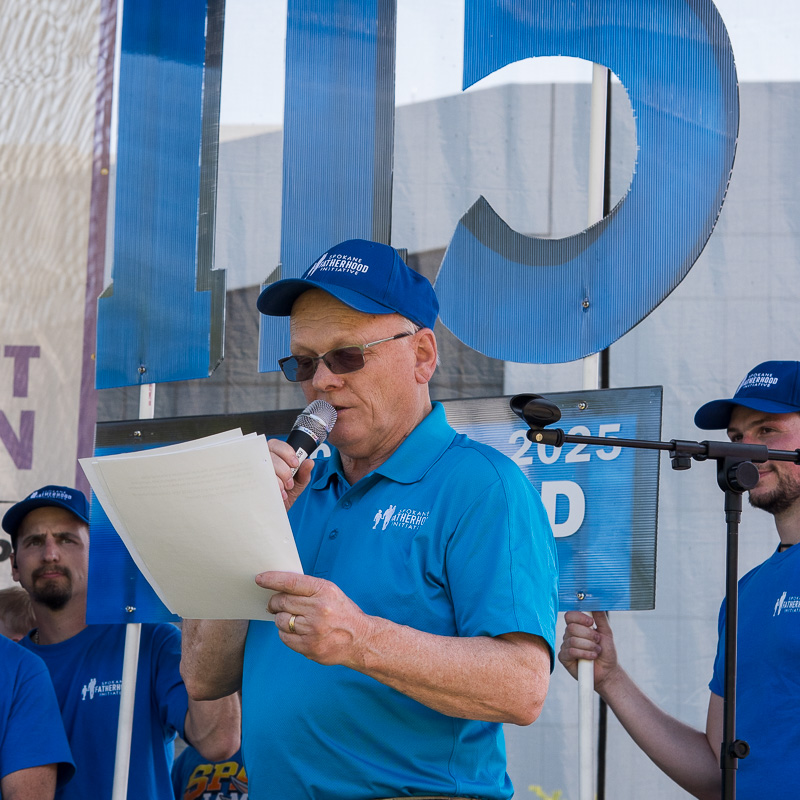 Community connection strengthens Spokane families at Father's Day celebration 12 Ron Hauenstein, SpoFI executive director, speaks into microphone while holding speech notes during Father of the Year announcement ceremony, with large 115th anniversary banner visible behind him