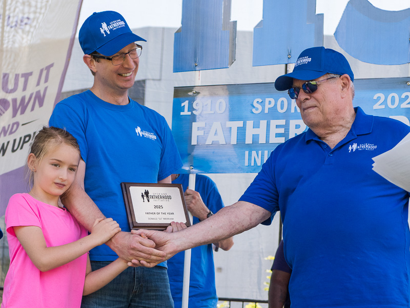 Community connection strengthens Spokane families at Father's Day celebration 13 DJ Merriam receives 2025 Custodial Father of the Year award plaque from Ron Hauenstein while his daughter in pink shirt helps hold the award, with 115th anniversary banner behind them