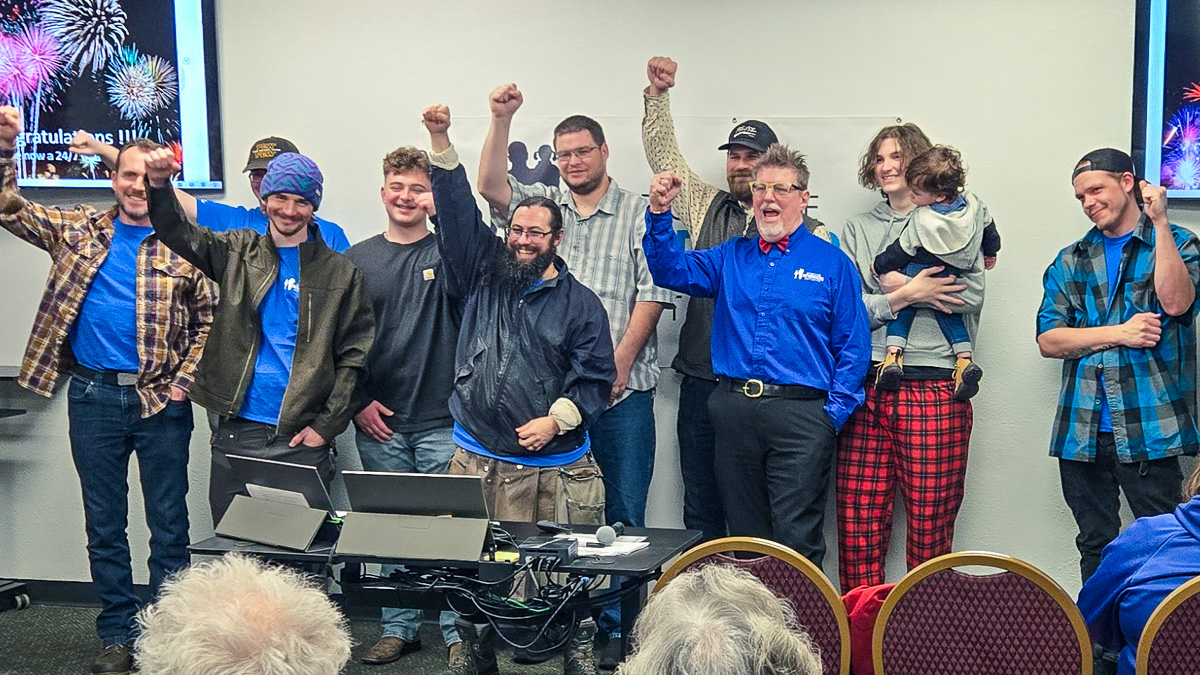 Proud graduates celebrating completion of SpoFI's 24/7 Dad parenting program in Spokane, raising their arms in triumph with instructors in blue SpoFI shirts. Group includes fathers of diverse ages gathered around a presentation table, some holding children, showing the supportive community spirit of the fatherhood classes.