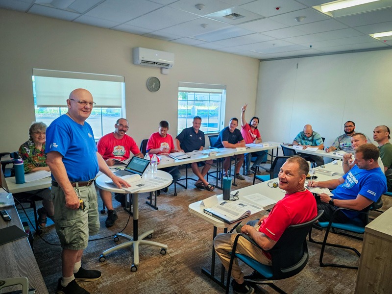 A man in a blue shirt leads a fatherhood class while a group of men sit at tables with workbooks in a well-lit classroom.