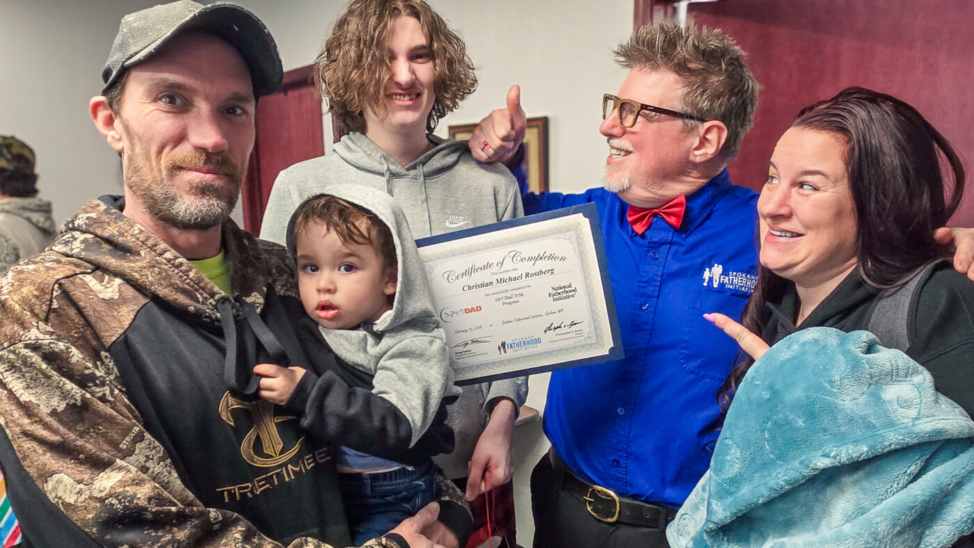 A proud family moment at SpoFI's 24/7 Dad® graduation - A young father in a camouflage jacket holds his toddler son, while standing with his teenage son in a grey hoodie, all beaming alongside a SpoFI instructor in a blue shirt with red bowtie and a woman in dark clothing. They're displaying a 24/7 Dad® completion certificate, capturing three generations celebrating this milestone together.